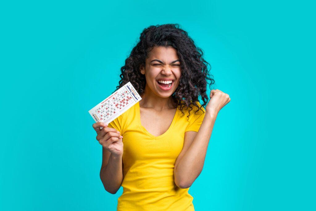 Eine glückliche Frau im gelben T-Shirt hält einen Lottoschein und jubelt vor Freude. Im Hintergrund ist eine türkisfarbene Wand zu sehen.