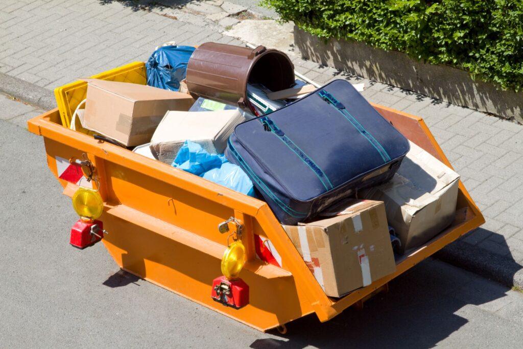 Ein orangefarbener Container, gefüllt mit verschiedenen Gegenständen wie Kisten, Taschen und Müll, steht am Straßenrand bereit zur Entsorgung im Rahmen einer Entrümpelung in Mönchengladbach.