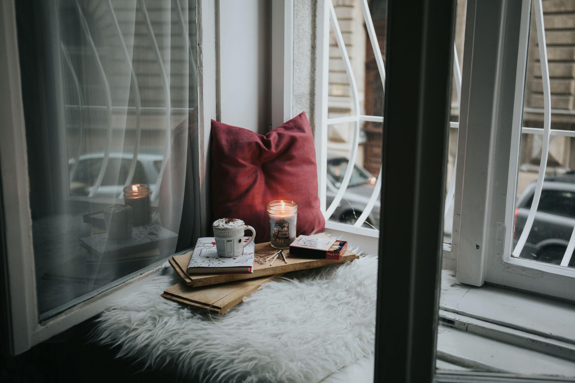 Fensterbank mit weichem Kissen und Kerze, zusammen mit Büchern und einer Tasse heiße Schokolade für eine gemütliche Atmosphäre