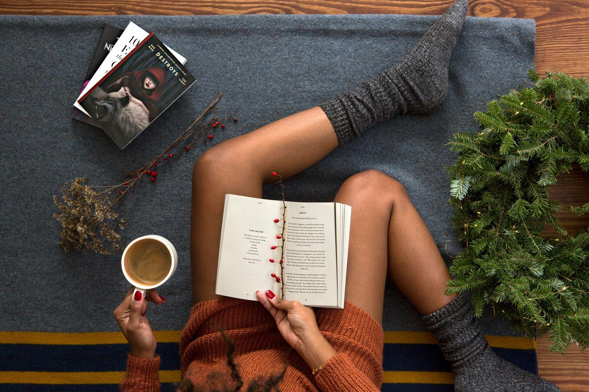 Person in gemütlichem Pullover sitzt auf einer Decke, liest ein Buch und hält eine Tasse Kaffee; umgeben von Tannenzweigen und Büchern